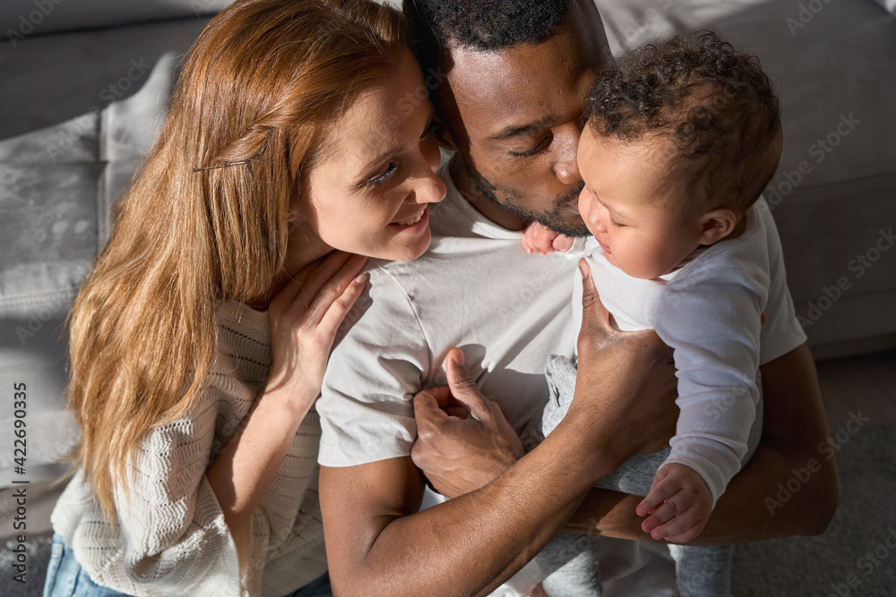 Mixed race young family couple bonding holding cute infant girl. Happy multiracial parents playing with small adorable baby daughter kissing kid at home lit with sunlight, Multiethnic family portrait Mixed race young family couple bonding holding cute infant girl. Happy multiracial parents playing with small adorable baby daughter kissing kid at home lit with sunlight, Multiethnic family portrait