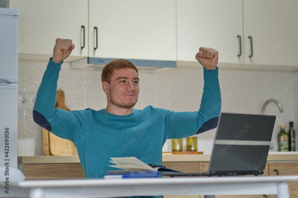 Happy excited man using laptop and raising his arms up to celebrate success at kitchen. success without leaving home, the new norm. Happy excited man using laptop and raising his arms up to celebrate success at kitchen. success without leaving home, the new norm.