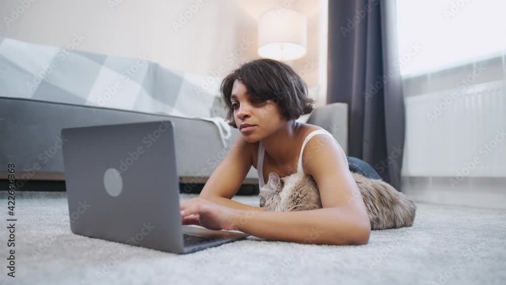 Young woman with a cat lies on the floor in the living room and works at a laptop, self-isolation at home during quarantine, remote work. Young woman with a cat lies on the floor in the living room and works at a laptop, self-isolation at home during quarantine, remote work.