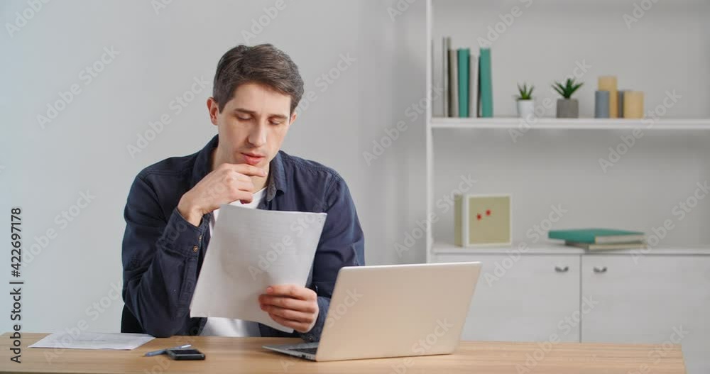Concentrated caucasian millennial businessman sitting at home desk or in office, looking at documents, reading letter paper, looking in financial statement, receiving notice from bank, doing paperwork Concentrated caucasian millennial businessman sitting at home desk or in office, looking at documents, reading letter paper, looking in financial statement, receiving notice from bank, doing paperwork