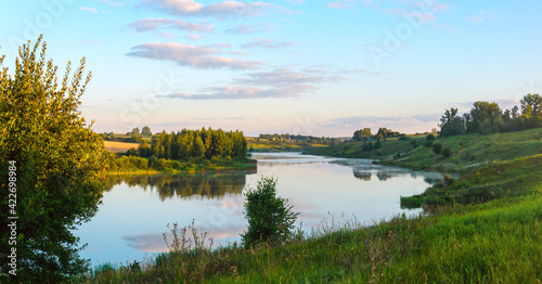 Summer serene landscape with calm river and green hills at sunrise.