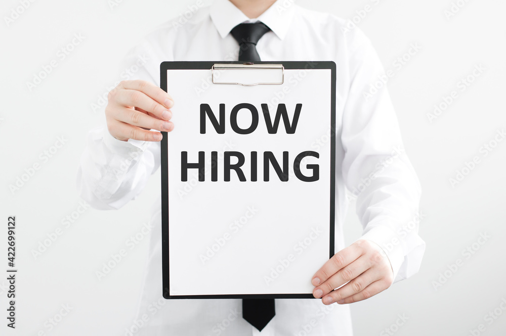 Now Hiring text on a white paper in the hands of a businessman, a man in a white shirt on a gray background. Now Hiring text on a white paper in the hands of a businessman, a man in a white shirt on a gray background.