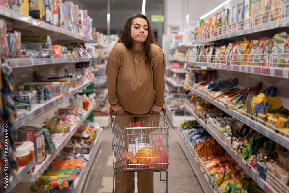 Caucasian female customer posing with shopping cart buying food in supermarket, pursing her lips. brunette choosing products in the store. .