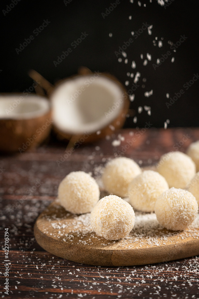 Coconut organic sweet truffles or homemade nut vegan energy balls with sprinkles of shredded coconut served on cutting board on dark brown wooden table at kitchen against black wall. Vertical Coconut organic sweet truffles or homemade nut vegan energy balls with sprinkles of shredded coconut served on cutting board on dark brown wooden table at kitchen against black wall. Vertical