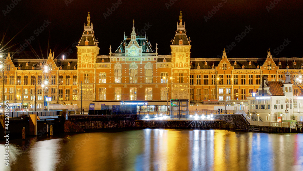 View of Central Station and the Amstel River with reflection of the lights in the water by night, Amsterdam, Netherlands.