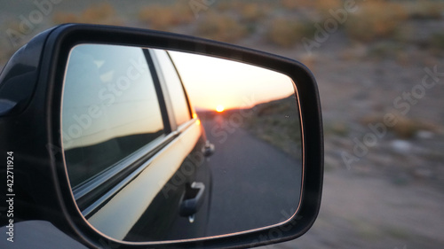 View in the side mirror of the car. Orange dawn over the hills. The car is going at high speed. Green fields, grass, and meadows are visible. Black color of the car.