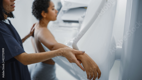 Friendly Female Doctor Explains the Mammogram Procedure to a Topless Latin Female Patient with Curly Hair Undergoing Mammography Scan. Healthy Female Does Cancer Prevention Routine in Hospital Room.