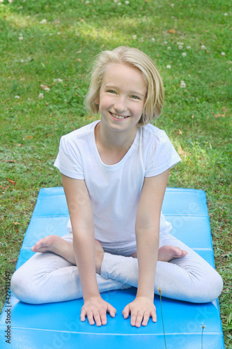 Beautiful and happy blonde kid girl of school age practices yoga on the mat in the park. Outdoor warm-up, healthy lifestyle. 
