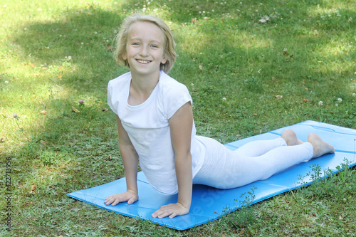 Beautiful and happy blonde kid girl of school age go in for sports and doing stretching on the mat in the park. Outdoor warm-up, healthy lifestyle. 