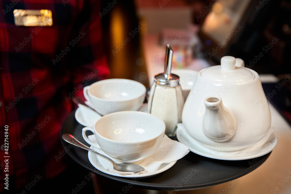 The waiter holds in his hand a tray with a kettle, tea cups and sugar. Close-up, selective focus. The waiter holds in his hand a tray with a kettle, tea cups and sugar. Close-up, selective focus.