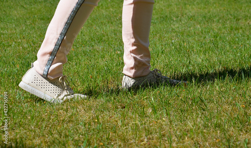 The feet of a girl in white sneakers on the green grass. Side view. A lady in athletic shoes walks in a green clearing. Summer, spring.
