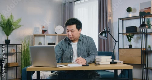 Good-looking confident hardworking adult asian man sitting at the table and working on computer in specially designeted place at home ,front view photo