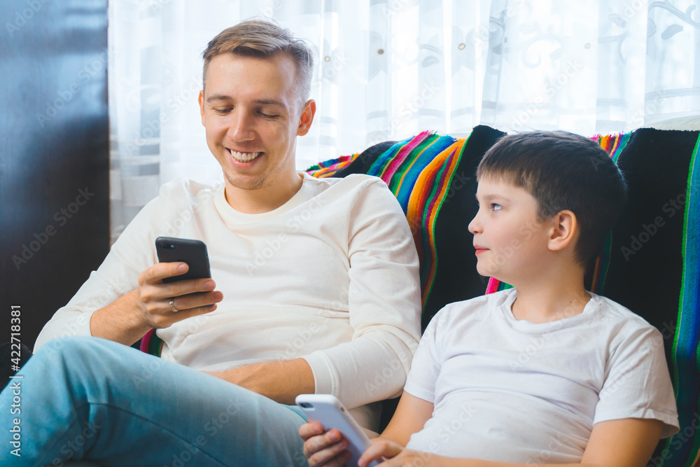 Cheerful young family dad son toddler childwatching online video on smartphone sitting on couch together, laughing parent kids texting playing games entertaining using mobile phone apps home Cheerful young family dad son toddler childwatching online video on smartphone sitting on couch together, laughing parent kids texting playing games entertaining using mobile phone apps home