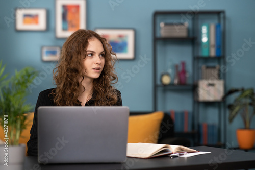 A young college girl is wondering what to write back to an email she received. The girl is sitting at a desk in a cozy living room using her laptop. The smiling teenage girl is studying remotely.