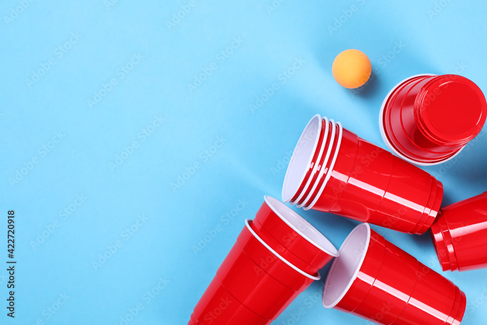 Plastic cups and ball on light blue background, flat lay with space for text. Beer pong game Plastic cups and ball on light blue background, flat lay with space for text. Beer pong game