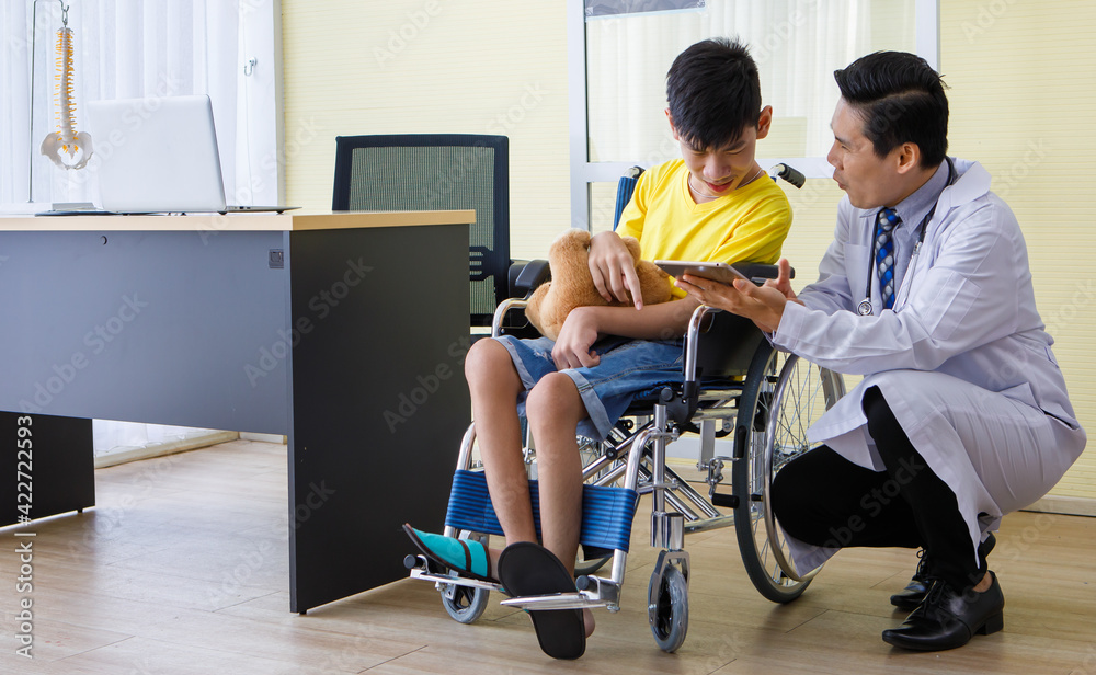 Asian doctor talking, support, and encouragement to a young disabled boy in a wheelchair. He shows tablet computer screen to the patient Asian doctor talking, support, and encouragement to a young disabled boy in a wheelchair. He shows tablet computer screen to the patient