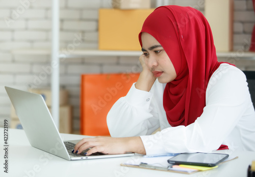 Portrait shot of young Muslim woman wearing a red hijab sitting on a black office chair, looking at the laptop with serious thinking about how to solve her company problems. Concept of stress at work
