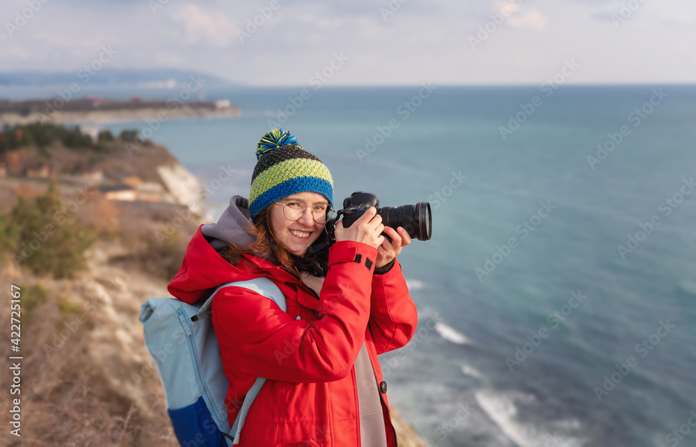 Young happy woman professional photographer in cap and red jacket traveling on the rocky seashore