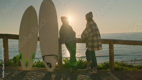 Two friends surfers stand on top of hill overlooking beach. Talk about waves and lineup during morning session. Beautiful sunrise or sunset light at popular surfing spot