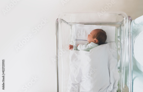 Newborn baby is sleeping in small transparent portable plastic bed. Baby first days of life is lying in a hospital crib after birth. photo