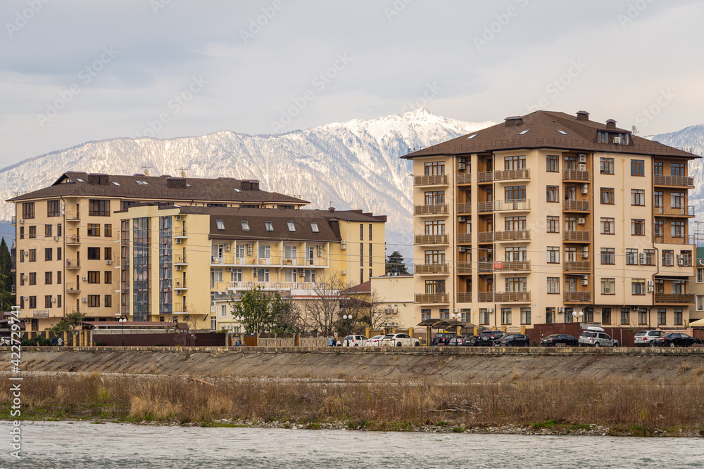 Residential buildings in federal territory of Sirius against backdrop of snow-capped peaks. Caucasus mountains. Imeretinskaya lowland. In foreground is Mzymta. Sirius, Russia - March 16, 2021