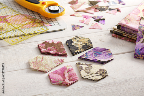 Stacks of multi-colored pentagonal pieces of fabric, ruler, cutting mat, rotary cutter on a white surface