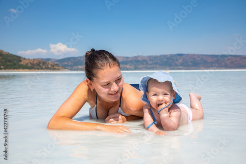 Young woman in swim suit and her daughter little baby girl are laying on white clay of turquoise crater lake Salda Golu, Turkey