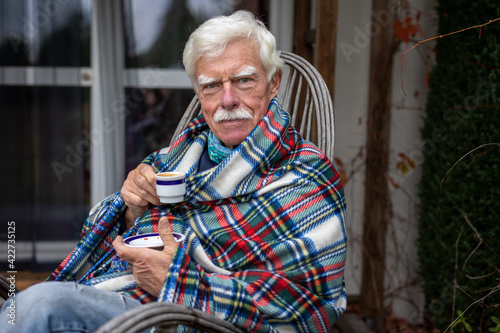 An older man wrapped in a blanket relaxes on a porch in the garden, drinking coffee.