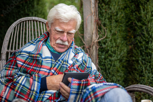 Senior man reading a book on his e-book reader when sitting in the rocking chair. Portrait of a happy, retired man reading a book on his device. 