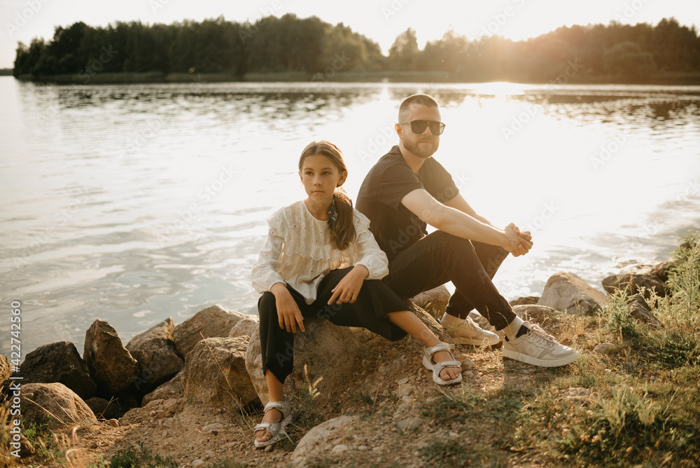 A young father with a beard and sunglasses is posing on stones with his daughter on the coast of the lake in the evening. Single-parent stylish family on vacation.