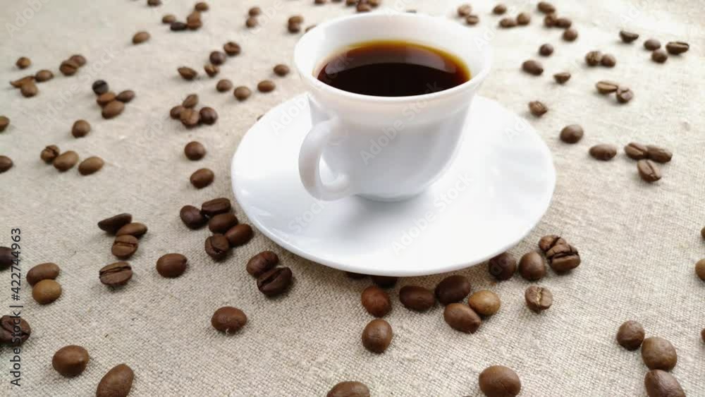 Espresso coffee in a small white cup on a light background of a textile napkin with scattered brown coffee beans. Breakfast concept, black coffee in a white cup. Selective focus, close-up