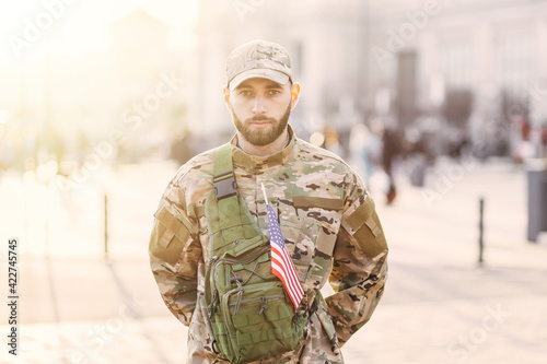 A young serious man in camouflage, in a cap with a backpack, stands on a city street.