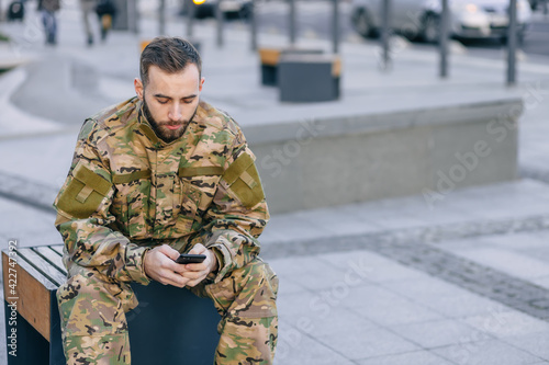 handsome male soldier in uniform sits on a park bench looking at the phone.