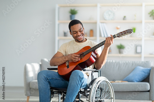 Handicapped black guy in wheelchair playing guitar and smiling at living room. Stay home activities