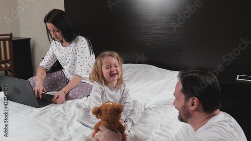 A young mother works at home on a laptop in the evening on the bed, the child and dad play paddle together. Happy family at home. Daughter and father play with toys, mother learns remotely online.