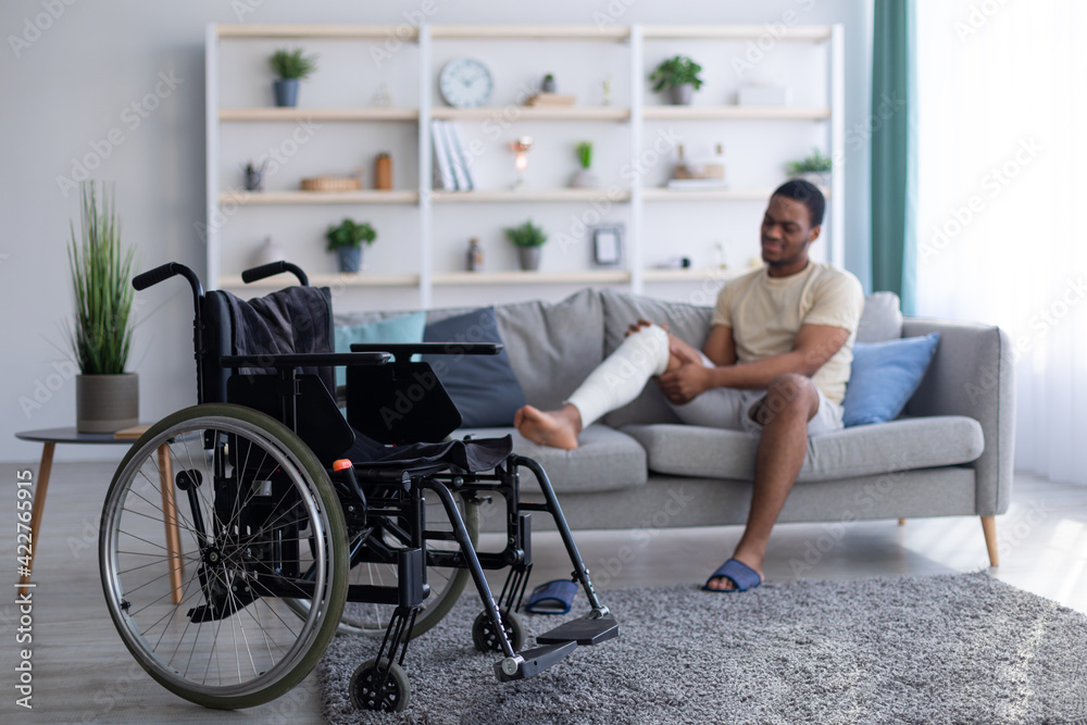 Young black guy with plastered broken leg sitting on sofa, focus on empty wheelchair, copy space