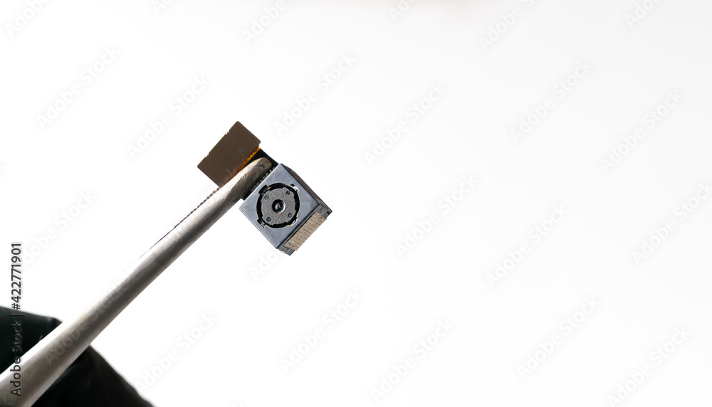 Close-up on the mobile phone camera module. Close-up tweezers or a clip holds the smartphone lens in a studio shot against a white background with copy space Close-up on the mobile phone camera module. Close-up tweezers or a clip holds the smartphone lens in a studio shot against a white background with copy space