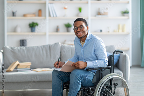 Fotobehang Happy disabled black guy in wheelchair taking notes, looking at camera and smili