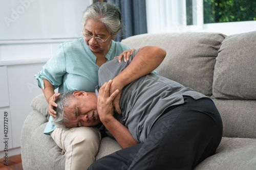 Asian old elderly man is In pain with his hands on his chest and having a heart attack in the Living room and his wife is shocked and confused, helpless. Concept of healthcare and Elderly caregivers © Prot