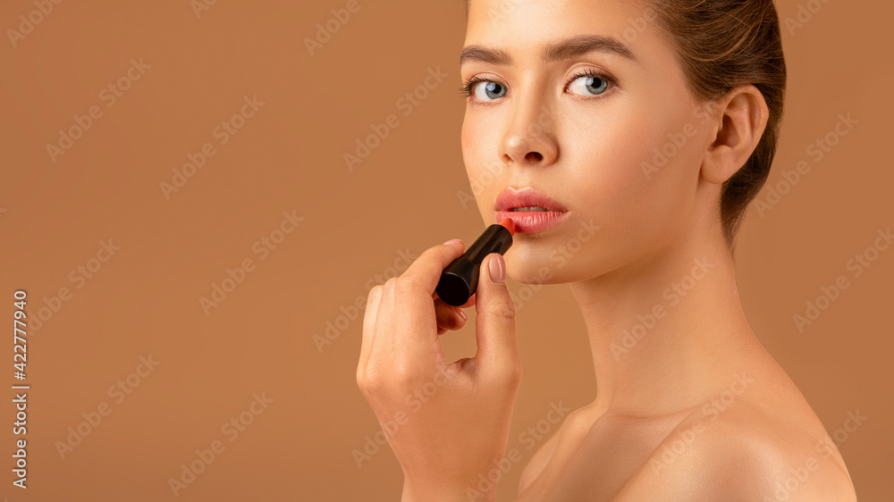 Lovely young lady applying lipstick, taking care of her appearance on brown background, panorama with copy space Lovely young lady applying lipstick, taking care of her appearance on brown background, panorama with copy space