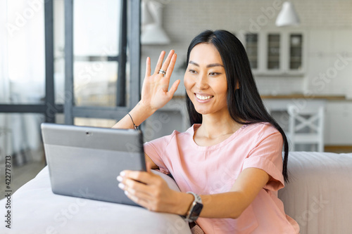 Cheerful young good looking Asian woman holding digital tablet and waving hand, talking via video call with friends or family, video conferencing, chatting on the pc sitting on the couch at home