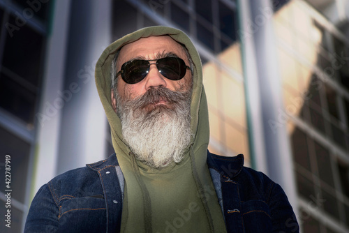 A senior man with a gray beard, wearing glasses from the sun, stands in front of a modern building. Casual clothing style photo