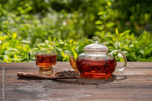 Steamed glass teapot, elegant cup. Bamboo spoon with tea, wooden table. Outdoor, picnic, brunch. Floral background in blur.