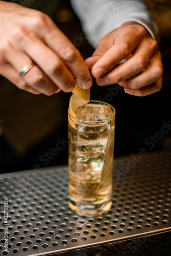 close-up of crystal glass with cold drink which barman s hands decorate with orange peel