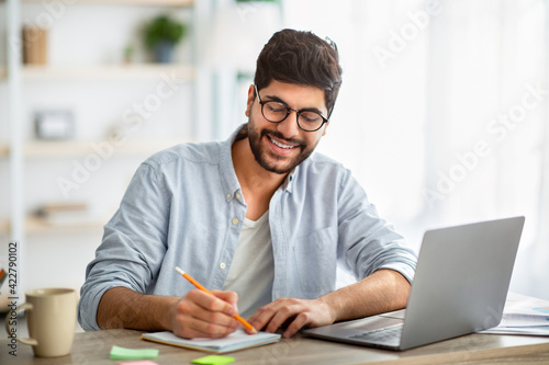 Happy arab man writing business report on laptop and taking notes, sitting at workplace at home interior