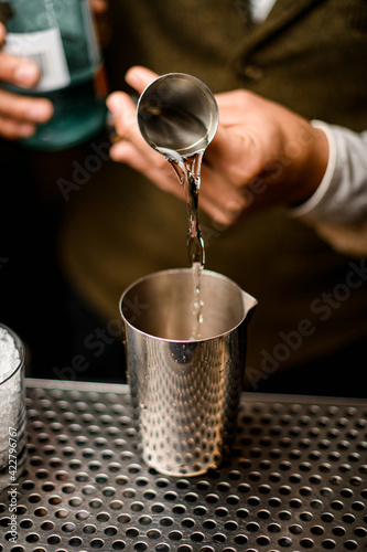 bartender holds steel jigger and accurate pours liquor into mixing cup on the bar