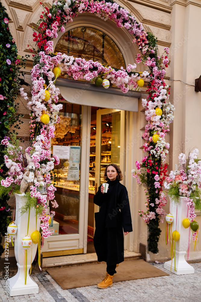 Beautiful young Caucasian woman near cafe, door decoration, Easter bunnies, artificial spring flowers, warm lights, lady, black coat and scarf, curly brown hair elegant glasses, romantic portrait