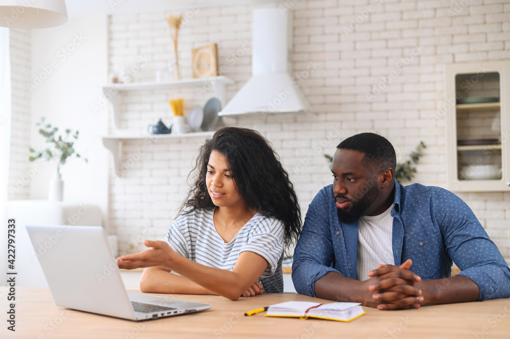 Two people african man and latin woman sitting on the kitchen at the laptop, study together online, discuss what they see, take notes, dressed in casual, making video call together, 4G connection Two people african man and latin woman sitting on the kitchen at the laptop, study together online, discuss what they see, take notes, dressed in casual, making video call together, 4G connection