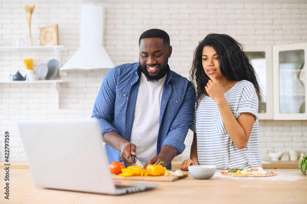 Happy african american spouses using laptop computer while cooking in modern kitchen together, searching recipe in internet, smiling black couple preparing healthy food for dinner at home, copy space
