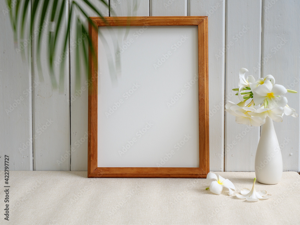 Mock up poster wooden frame and beautiful Frangipani tropical flower in modern white vase on beige table and wood wall background with green palm leaf